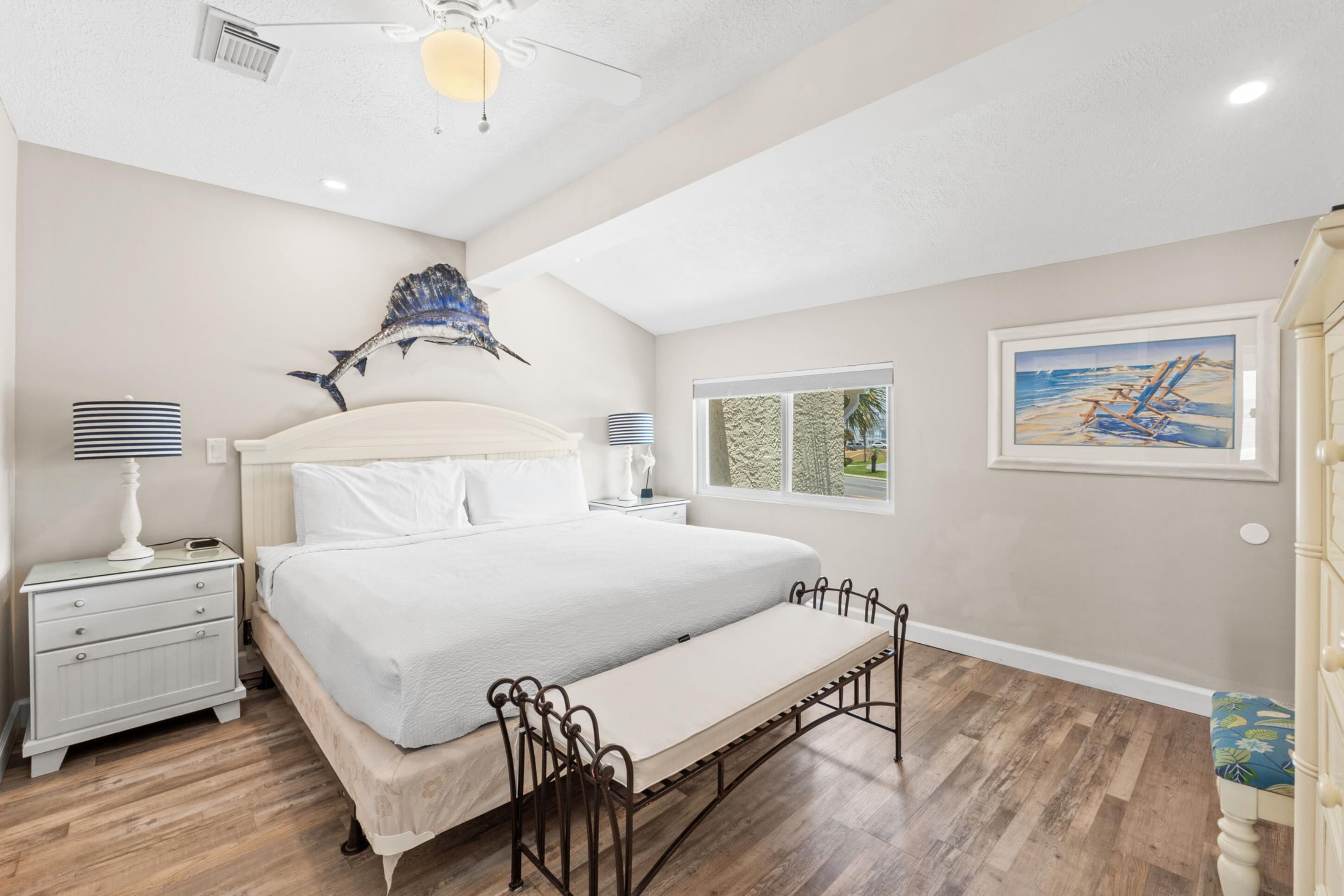 A bedroom with a bed, bedside tables, a bench, a ceiling fan, and artwork on the walls.