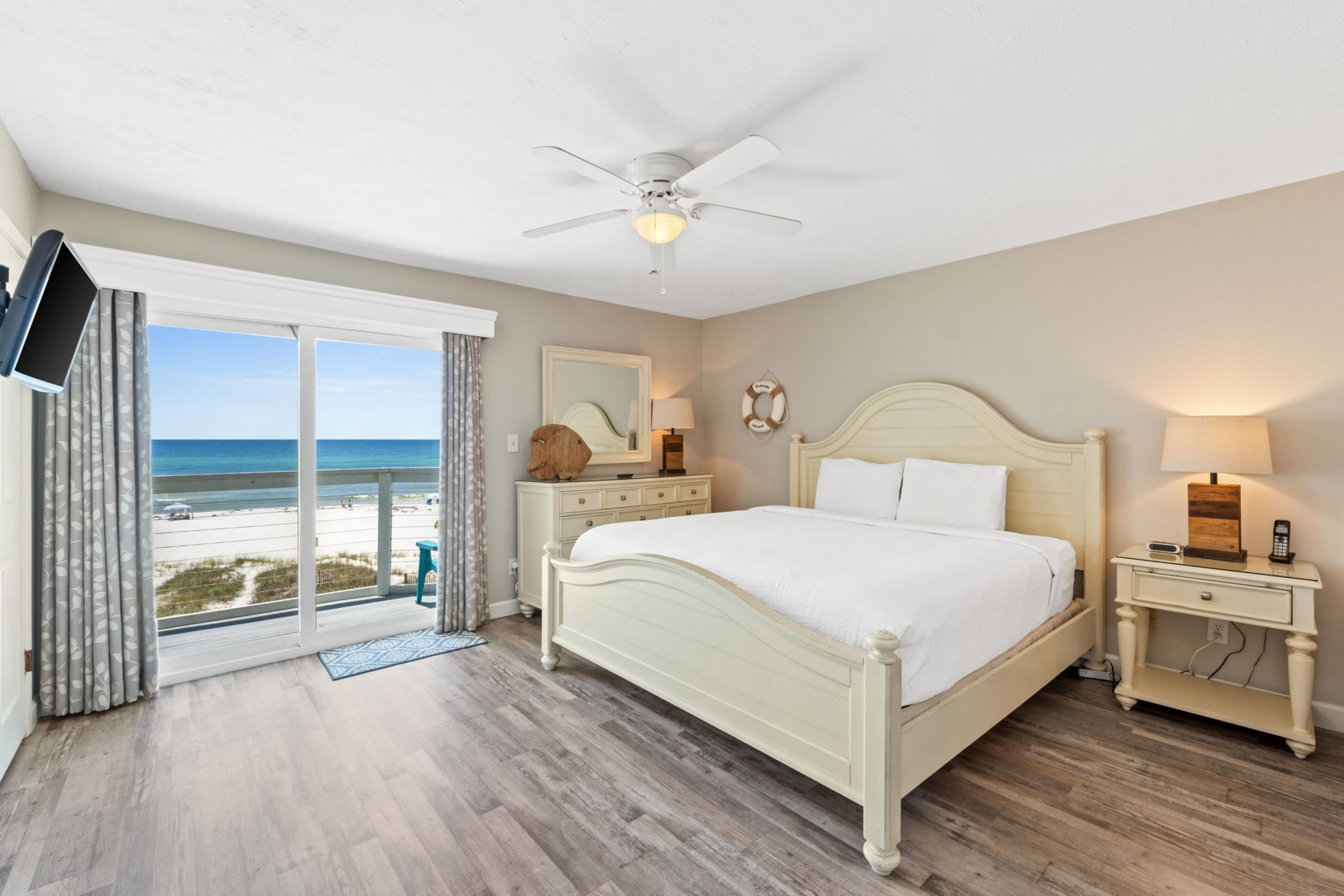 Une chambre à coucher comprenant un lit, des tables de chevet, un ventilateur de plafond, un téléviseur fixé au mur et une vue sur la plage.
