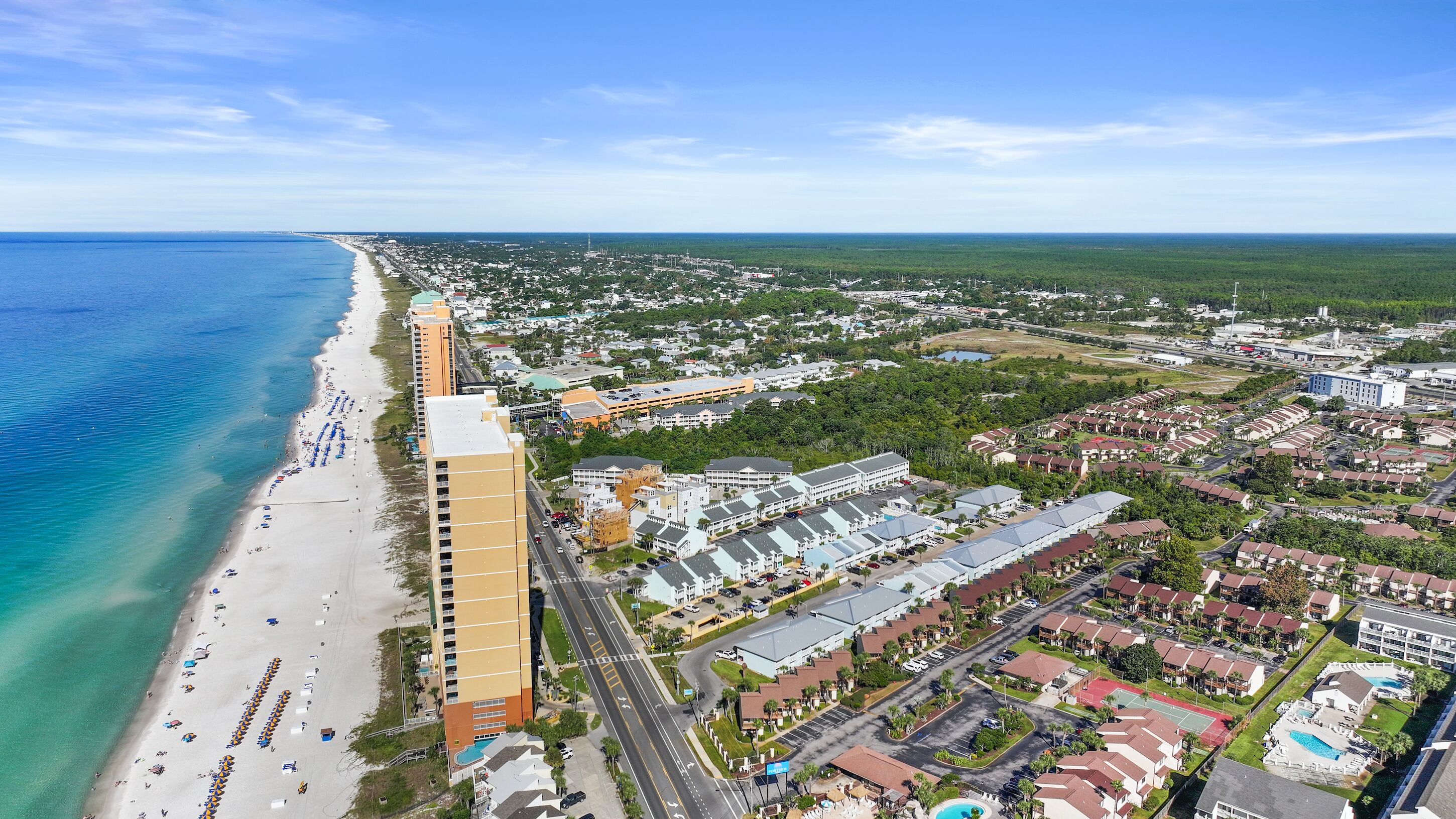 An aerial view of a coastal town with a long sandy beach, a large hotel with multiple buildings, and a mix of residential and commercial areas.