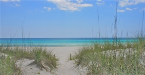 A beach with sand and sparse grass in the foreground, leading to a calm sea under a clear sky. - Southwind Condo Rentals by Panhandle Getaways (Panama City Beach)