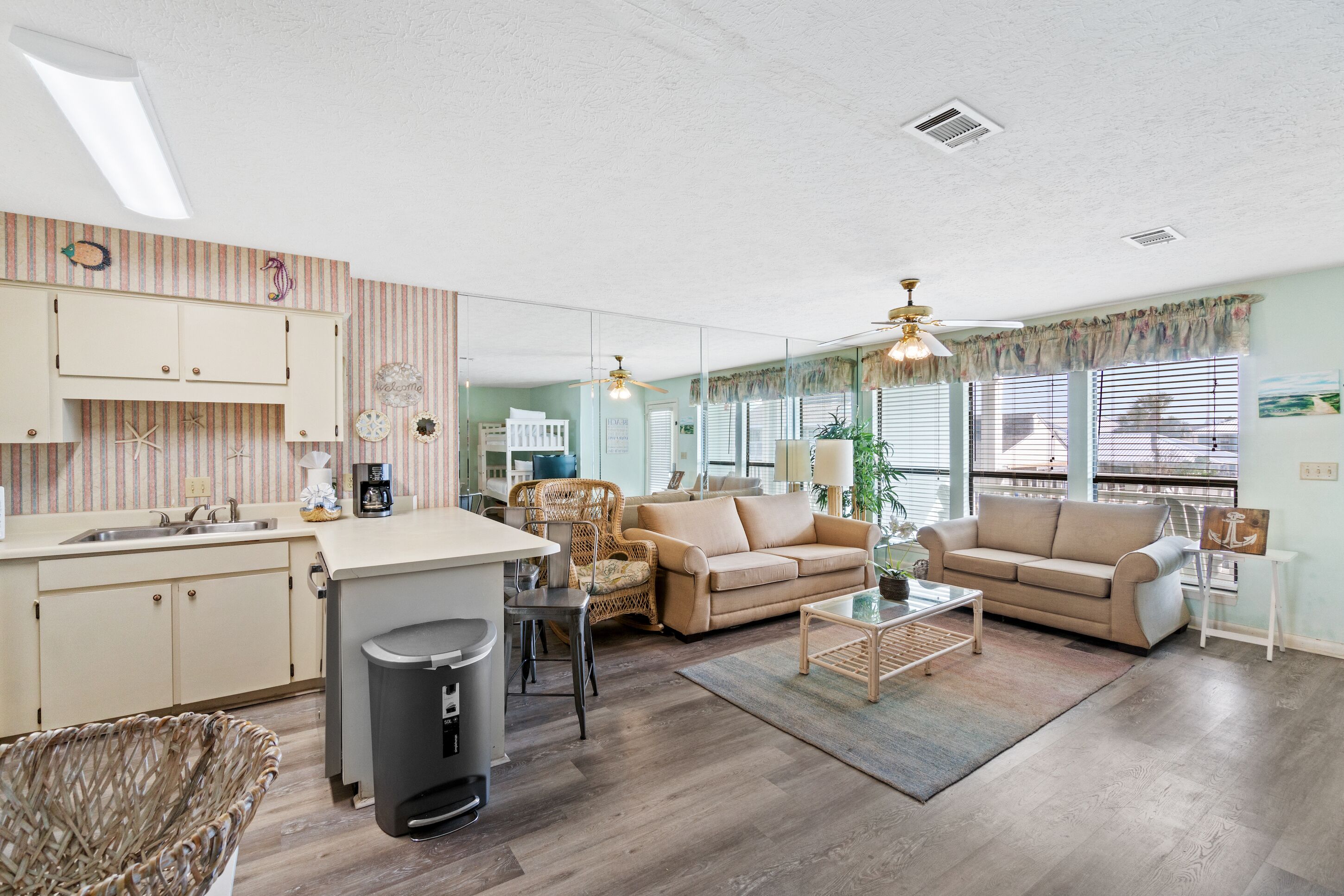Un vaste espace de vie comprenant une cuisine, un canapé beige, un fauteuil en osier et une table basse.