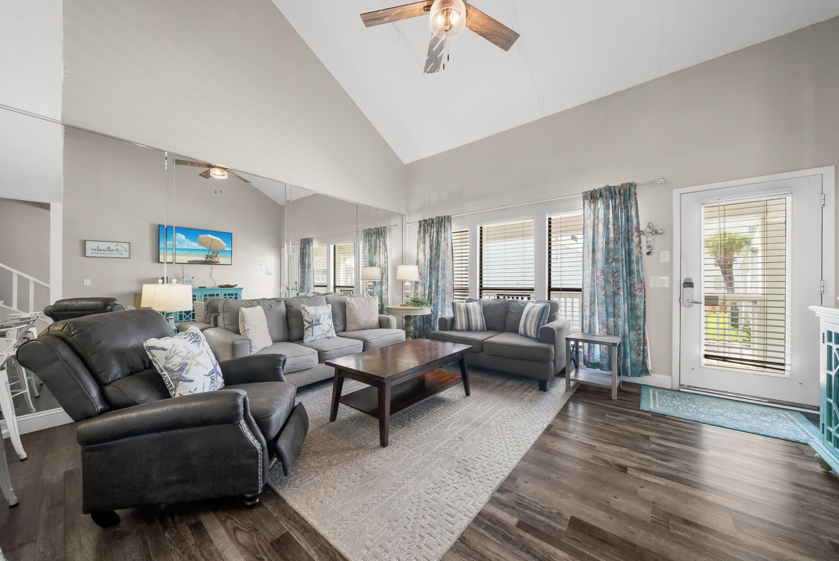 A spacious living room with a high ceiling, a ceiling fan, and a large window with blinds. There are multiple sofas and a coffee table.