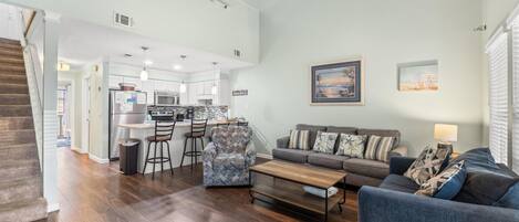 A spacious living area with a sofa, coffee table, and patterned armchair. A kitchen is visible in the background with a dining area and a staircase leading to another level.