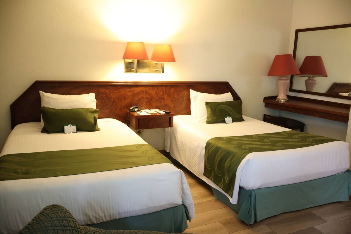 Una habitación de hotel con dos camas individuales, una mesita de noche, un escritorio con lámpara y un espejo.