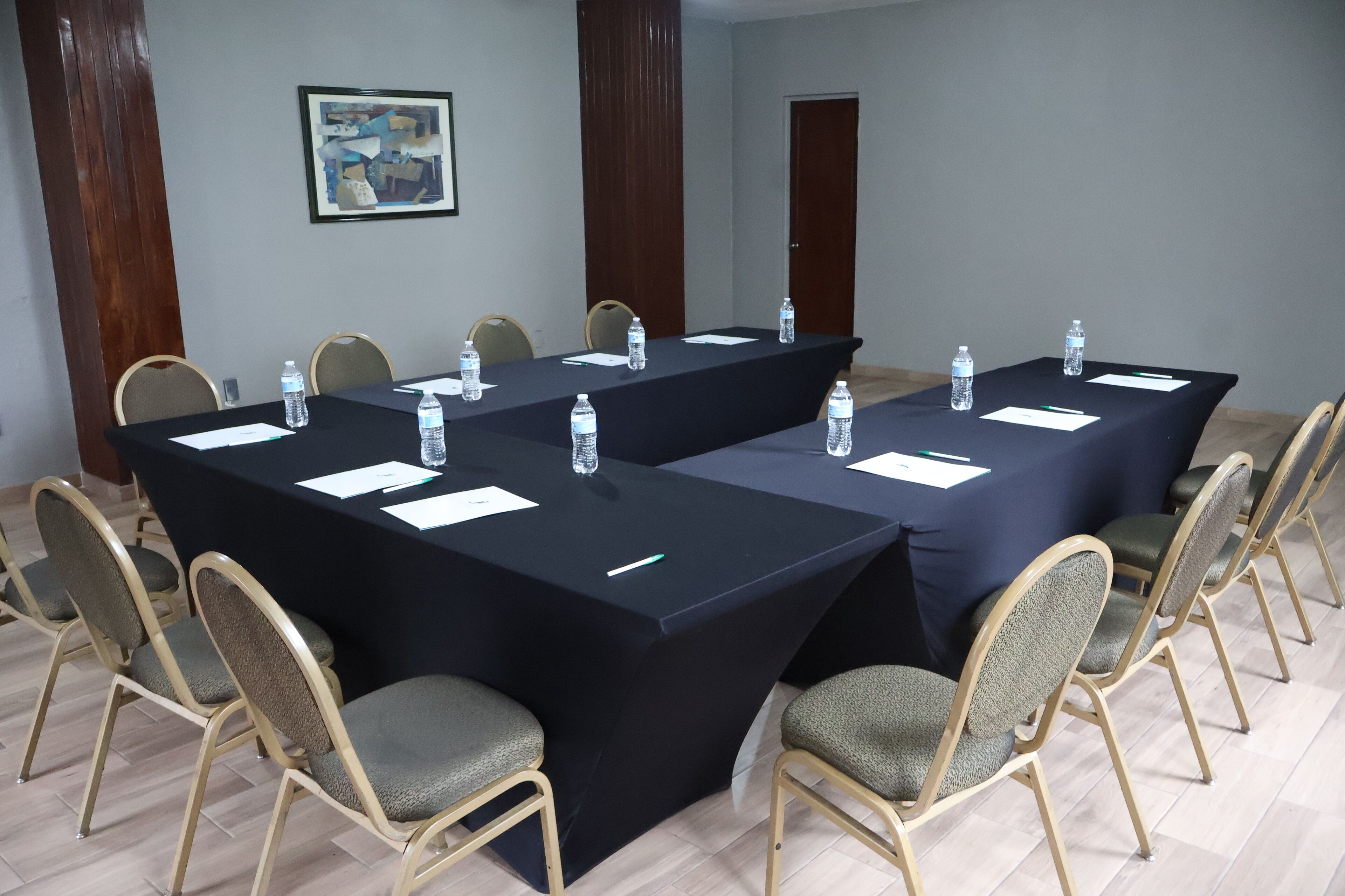A conference room with two long tables, each surrounded by six chairs. Water bottles and name tags are placed on the tables.