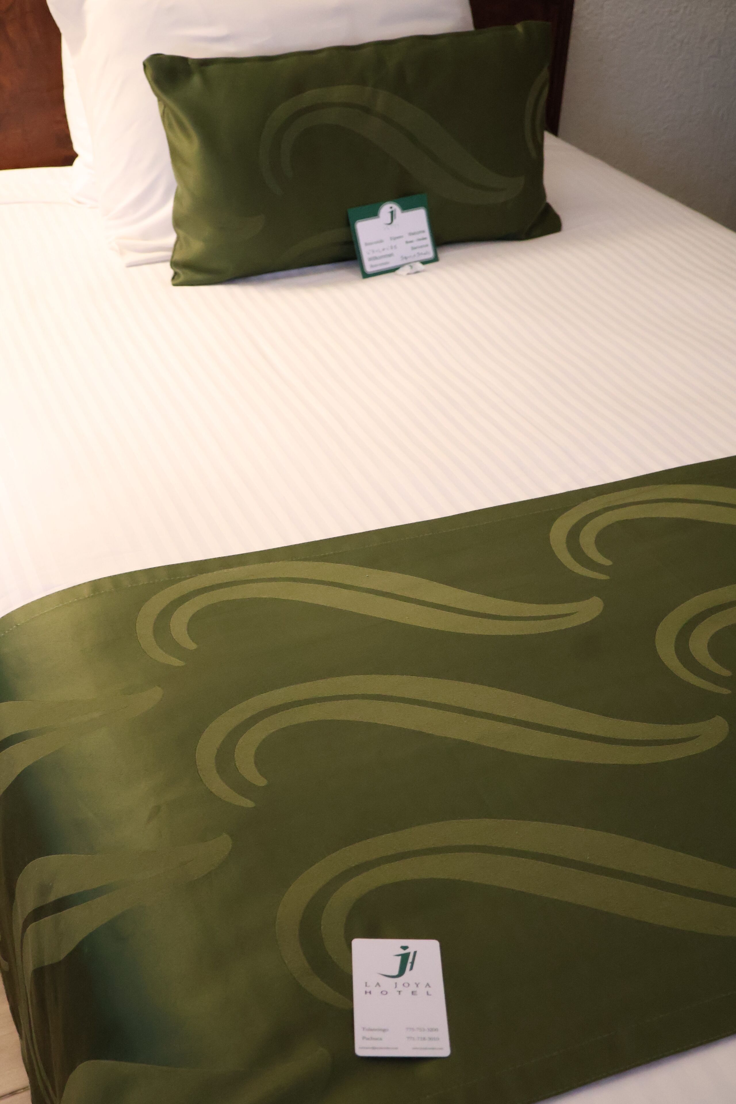 Una cama de hotel con una colcha verde y un almohadón, una etiqueta en la colcha, y un almohadilla blanca sobre la cama.