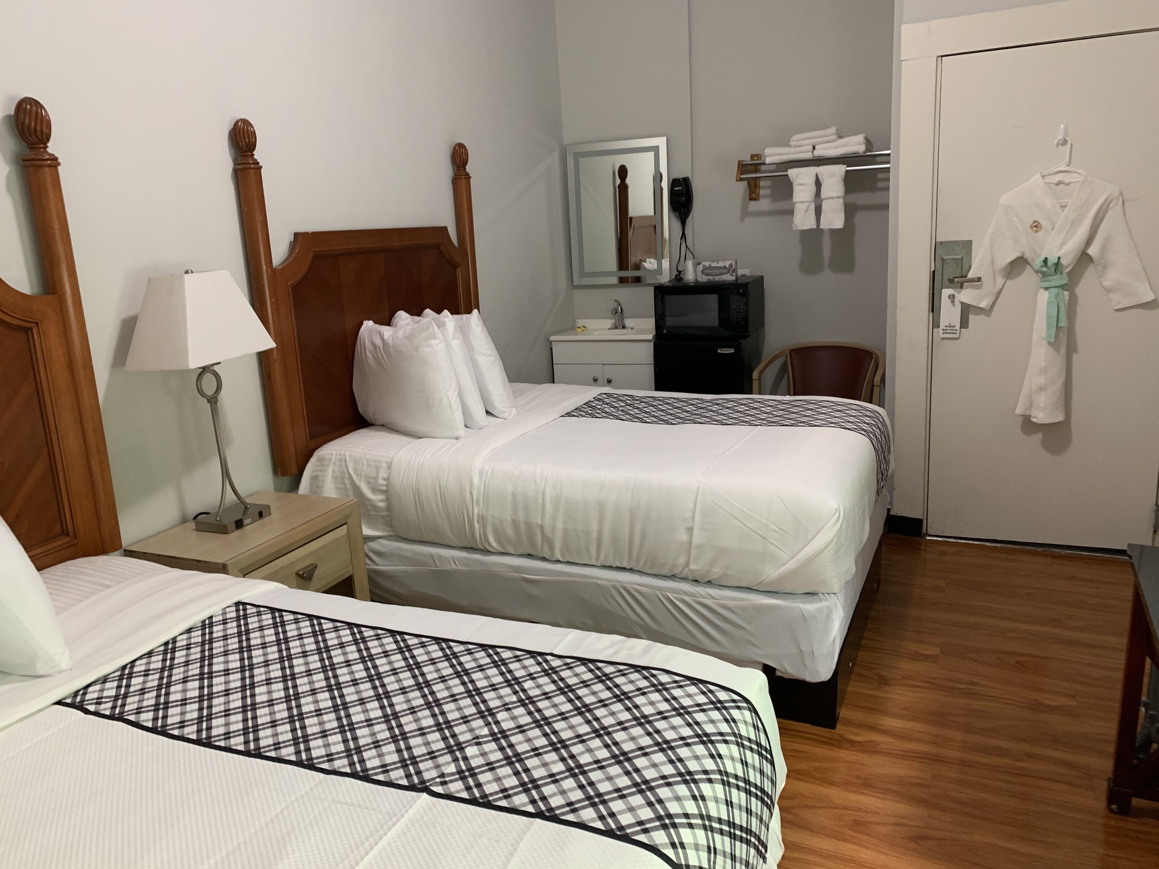 A hotel room with two beds, a bedside table with a lamp, a dresser, a mirror, a microwave, and a door with towels and robes hanging.