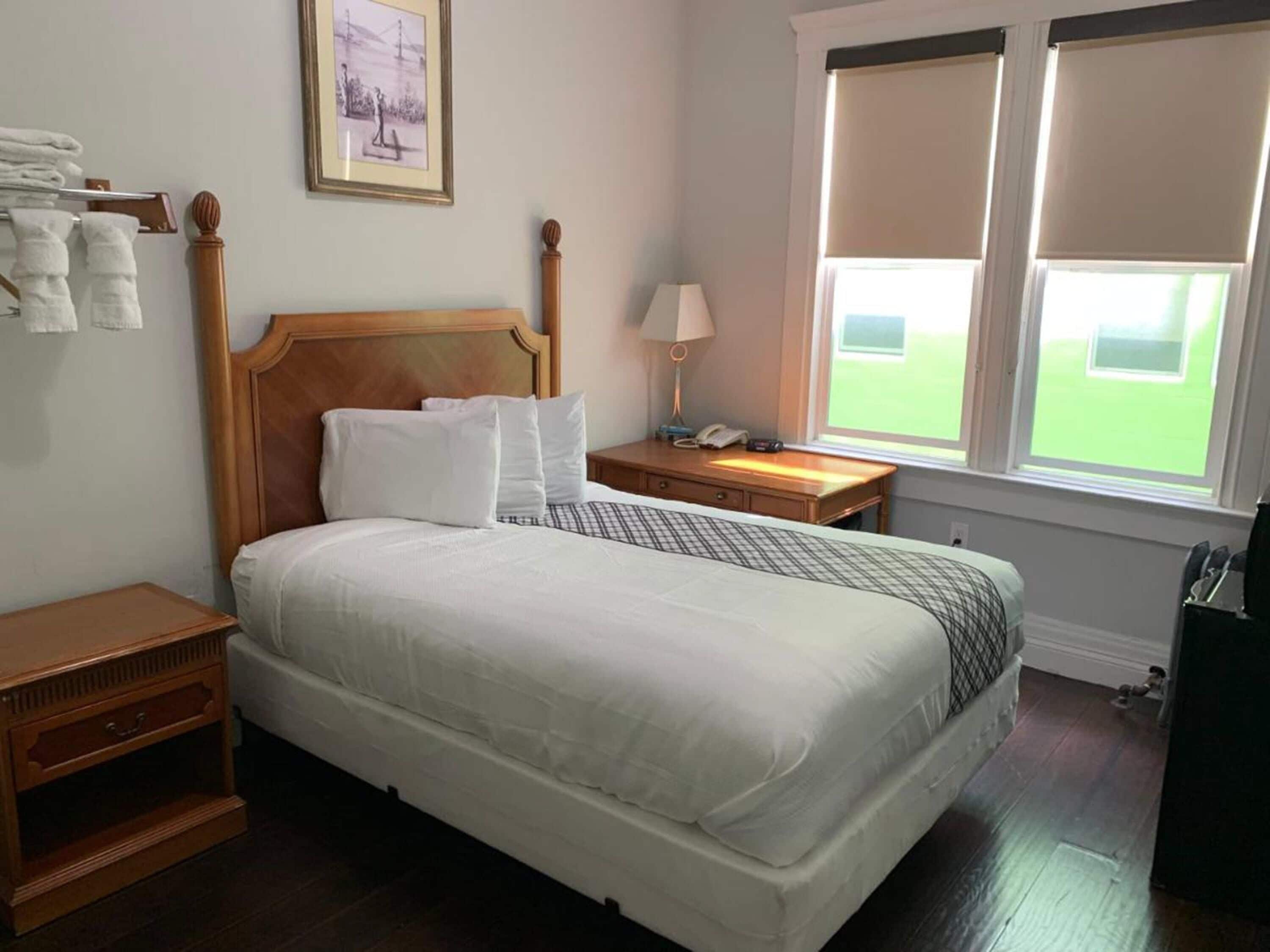 A neatly made bed with white linens, a wooden headboard, a bedside table with a lamp, and a window with blinds.