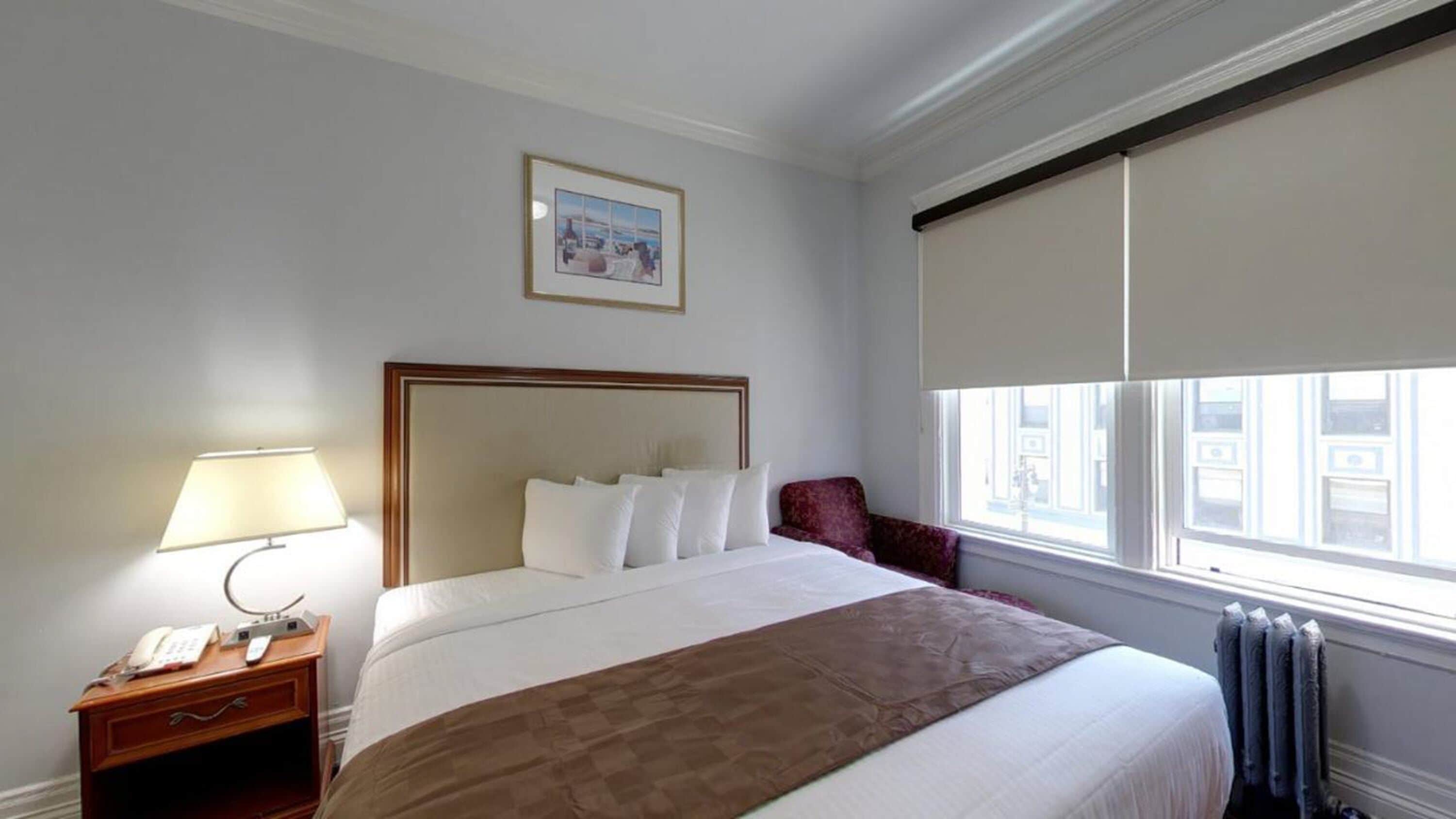 A hotel room with a large bed, a bedside table with a lamp, a chair, a window with blinds, and a framed picture on the wall.