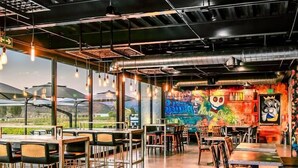 Um espaço de refeições interno com mesas e cadeiras, um mural na parede e vista para o exterior.