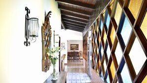 Um corredor com piso de azulejos, uma mesa de madeira com um vaso de flores, uma luminária na parede e uma parede estampada com janelas em formato de losango.