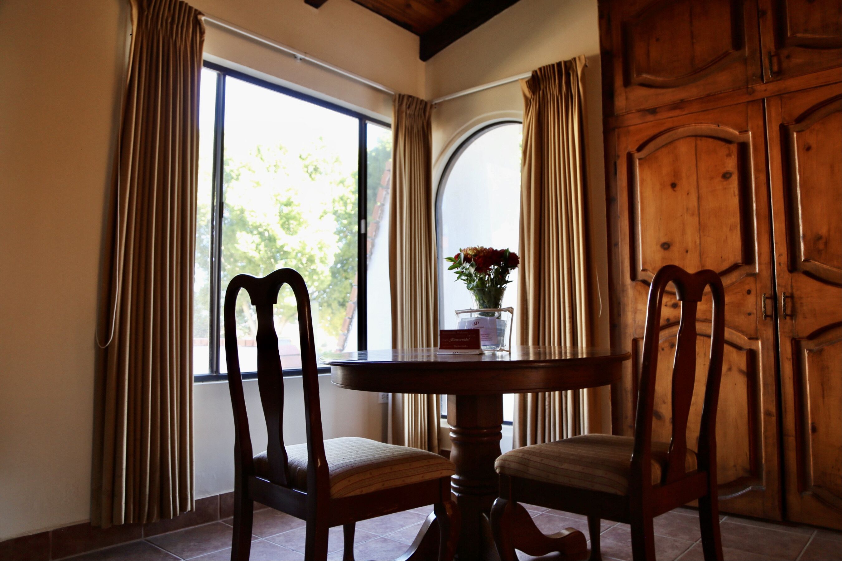 A dining area with a round table and two chairs, a vase with flowers, and a wooden door in the background.