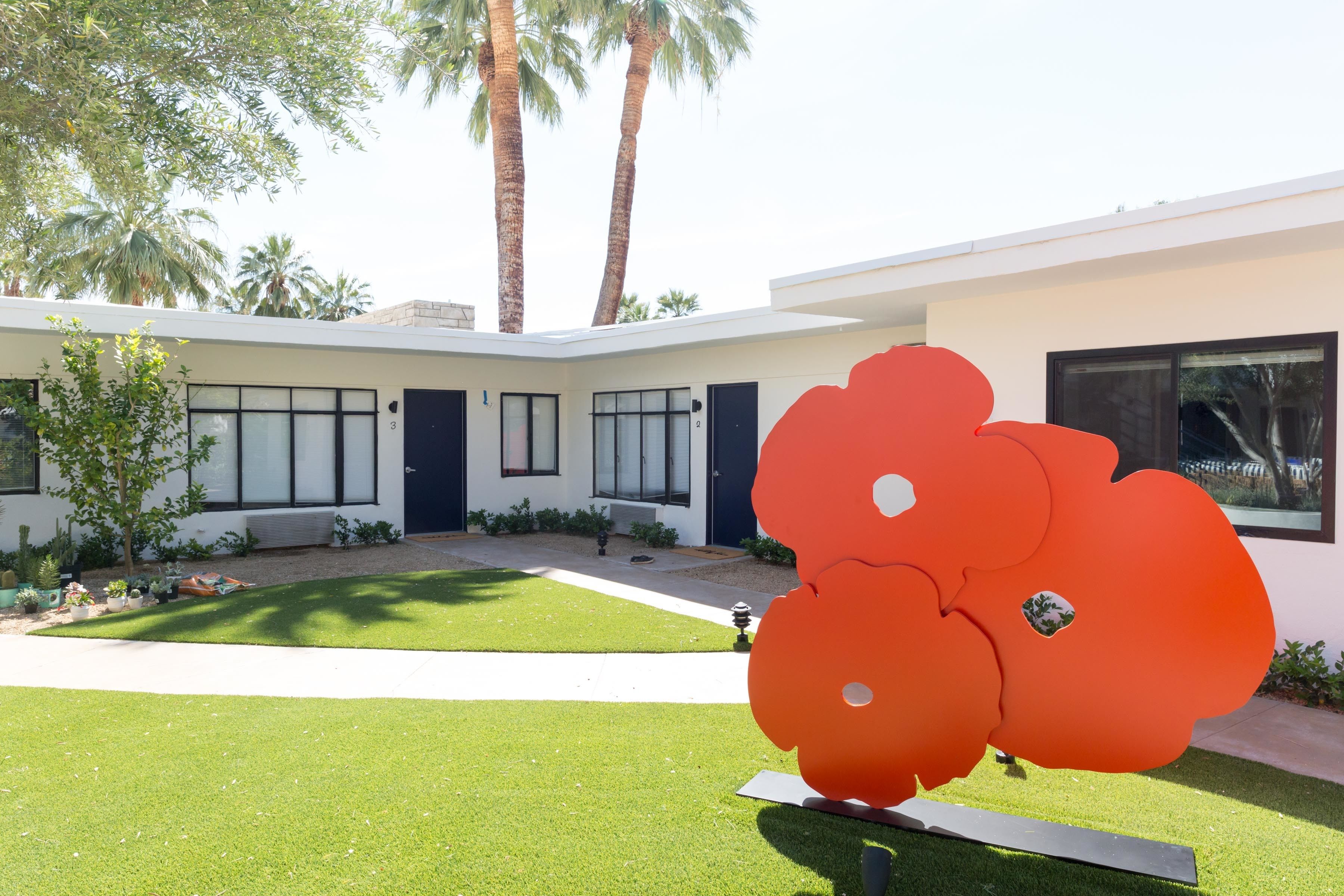A modern building with a well-maintained lawn, a large red sculpture, and palm trees in the background.