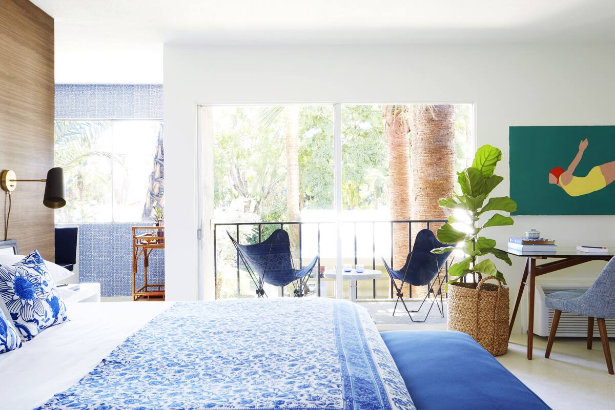 A bedroom with a large bed, a balcony with chairs, a dining table, and a potted plant.