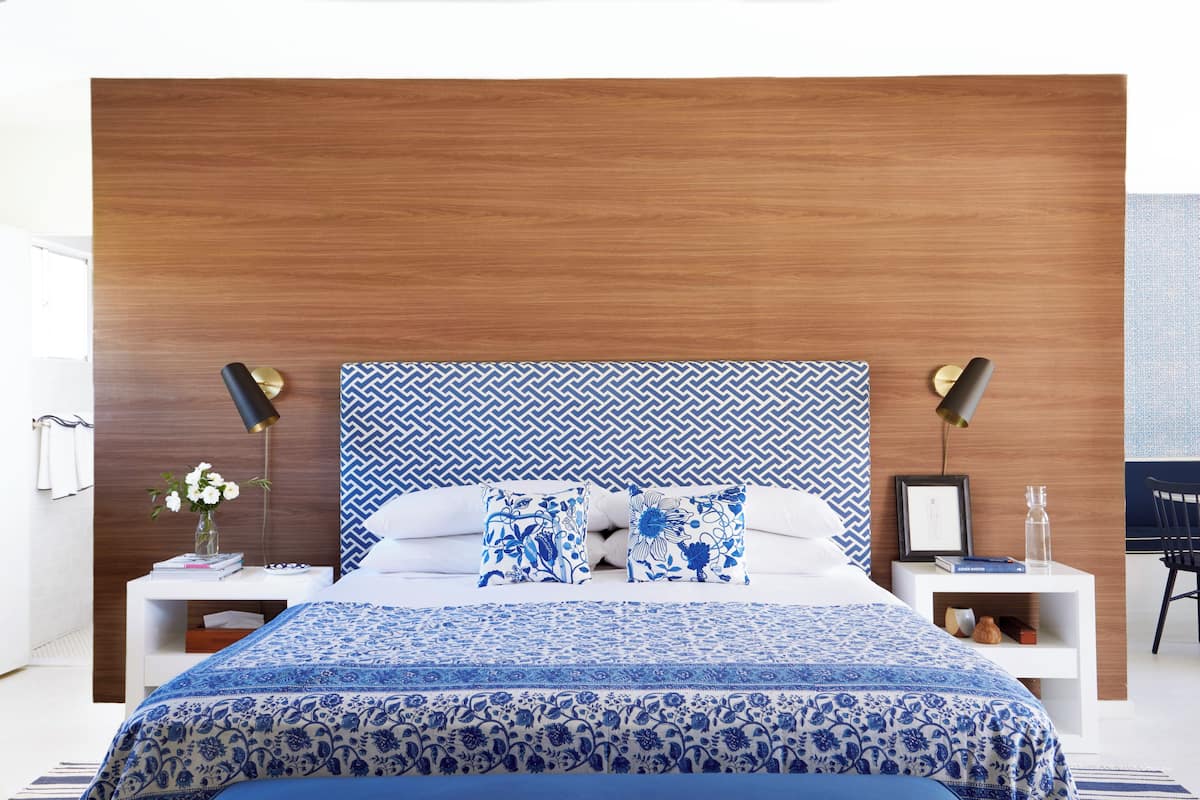 A modern bedroom with a wooden headboard, a patterned blue bedspread, and matching pillows. There are bedside tables with lamps and a small shelf with a book and a bottle.