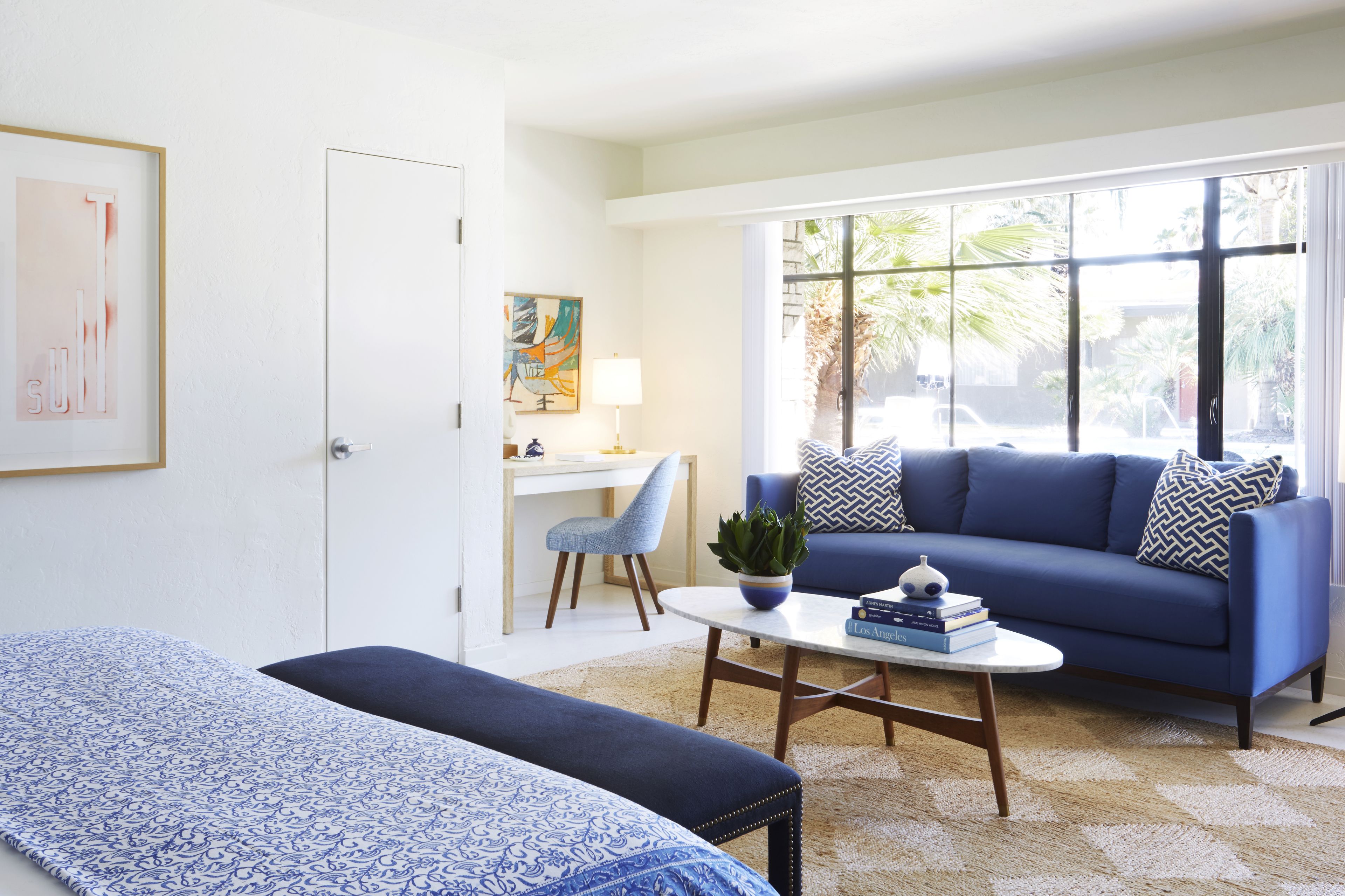 Un salon moderne avec un canapé bleu, une petite table basse et un bureau avec une chaise. Une grande fenêtre offre une vue sur les palmiers.