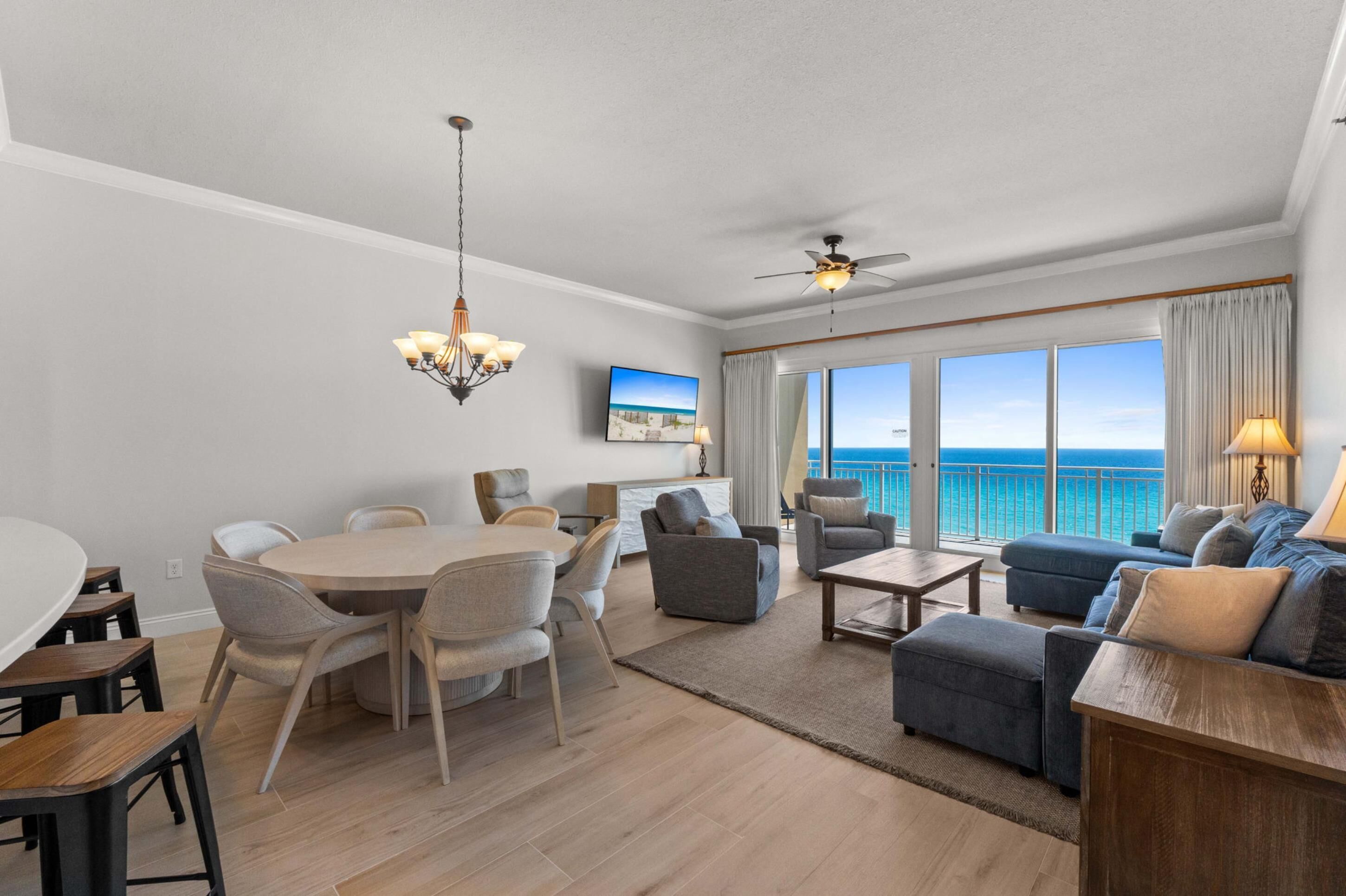 A modern living room with a dining area, large windows with a view of the ocean, a ceiling fan, and a chandelier.