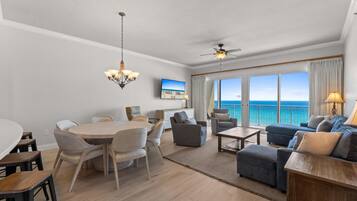 A modern living room with a dining area, large windows with a view of the ocean, a ceiling fan, and a chandelier.