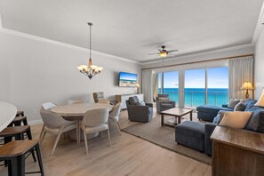 A modern living room with a dining area, large windows with a view of the ocean, a ceiling fan, and a chandelier.