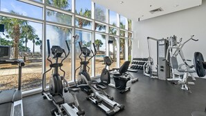 Un gimnasio con diversos equipos de ejercicio, amplias ventanas y vistas a palmeras.