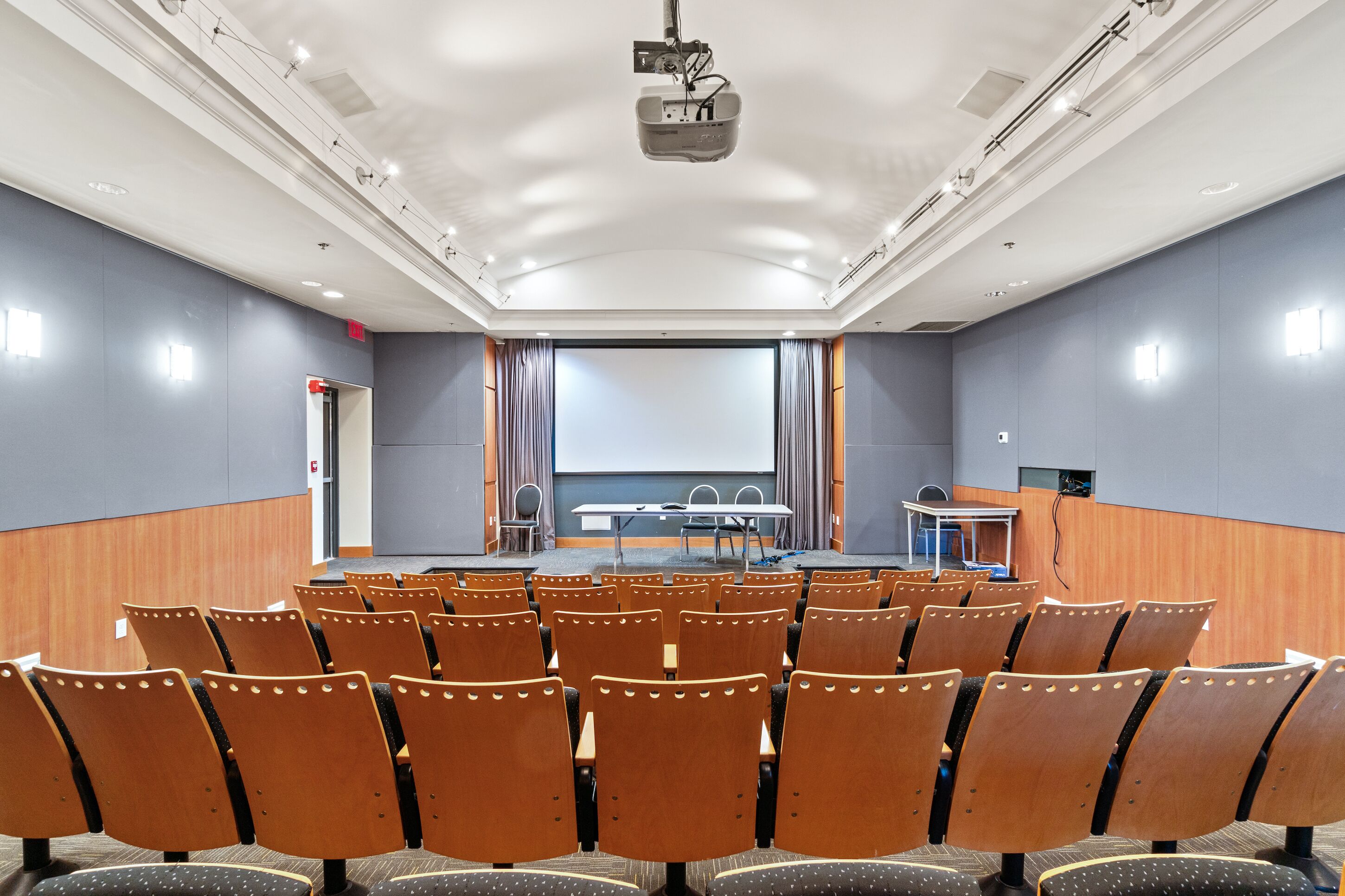 A conference room with rows of wooden chairs, a large screen, a table with chairs, and a podium.