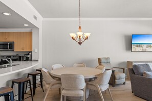 A modern kitchen with a central island, a dining area with a round table and chairs, and a living area with a sofa and TV.