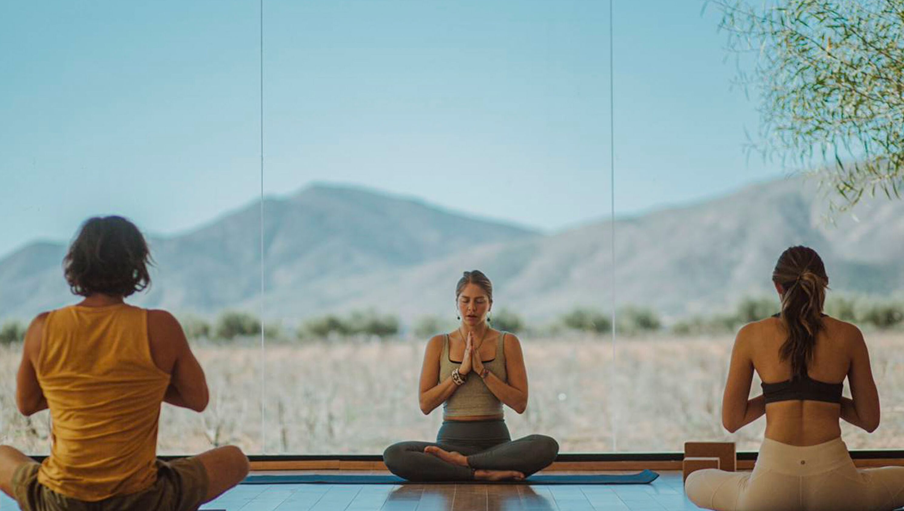Three individuals seated in a meditative pose on a yoga mat in a room with a large window overlooking a mountainous landscape.