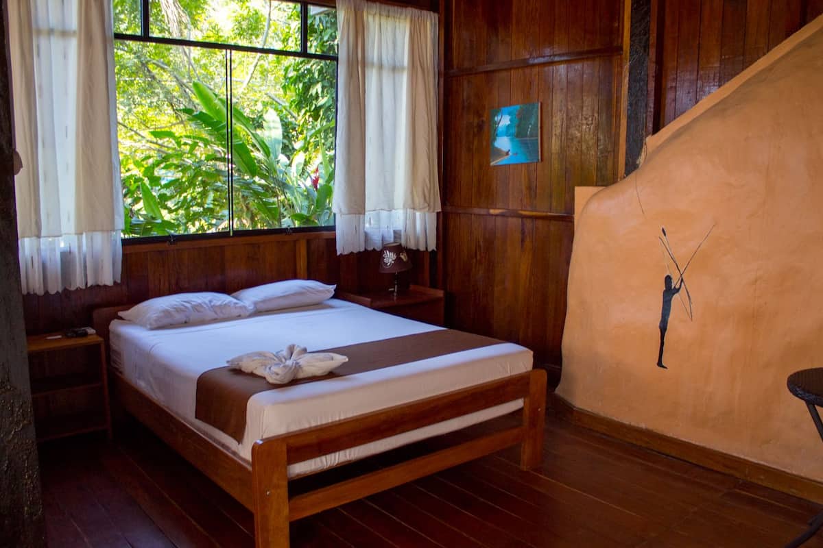 A wooden bed with white linens, a nightstand, a bedside lamp, and a view of greenery through a window with white curtains.