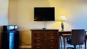 Habitación de hotel con televisión de pantalla plana montada en la pared, cómoda de madera, escritorio con lámpara y una silla.