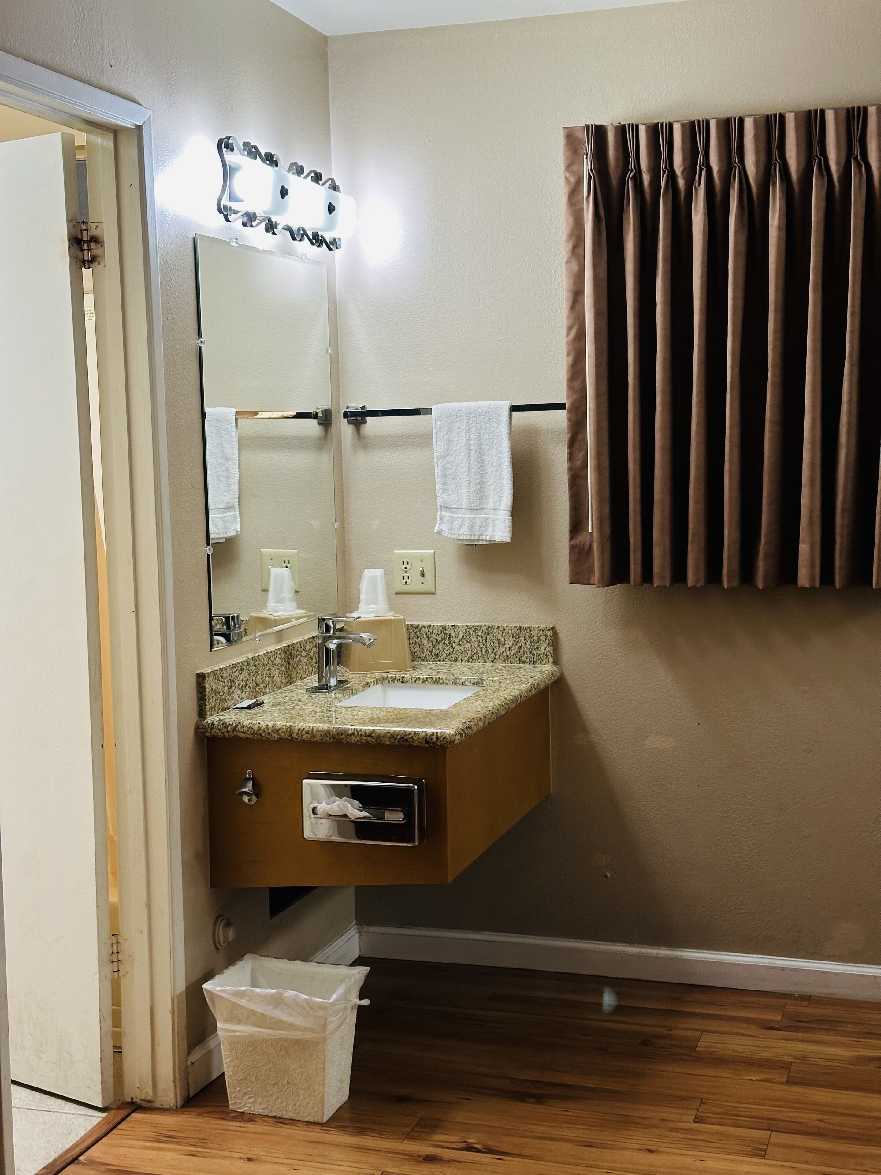 A bathroom with a granite countertop sink, a mirror with lights, a towel rack, and a wooden floor.