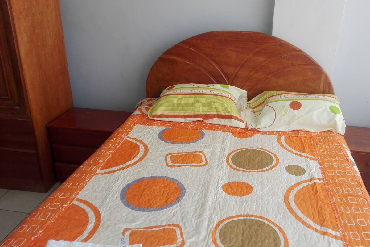 Una cama con un edredón estampado y dos almohadas, un cabecero de madera y una mesita de noche con un cajón.