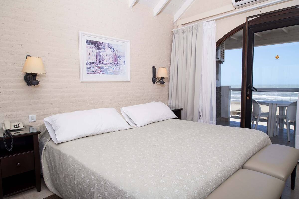 A hotel room with a bed, two bedside lamps, a phone, a framed picture, and a balcony with a view of the sea.
