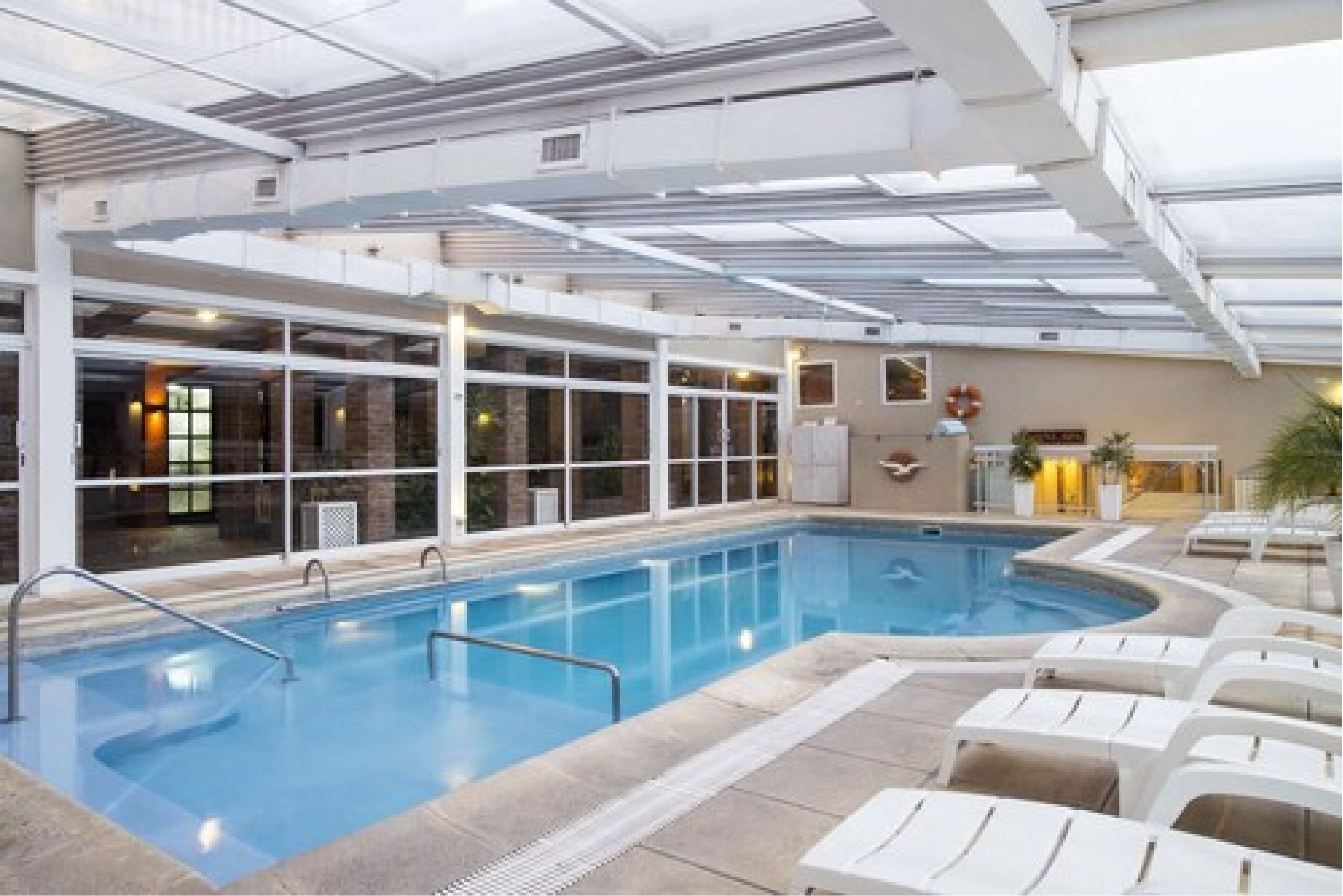An indoor swimming pool with clear glass walls, lounge chairs, and a ceiling with skylights.