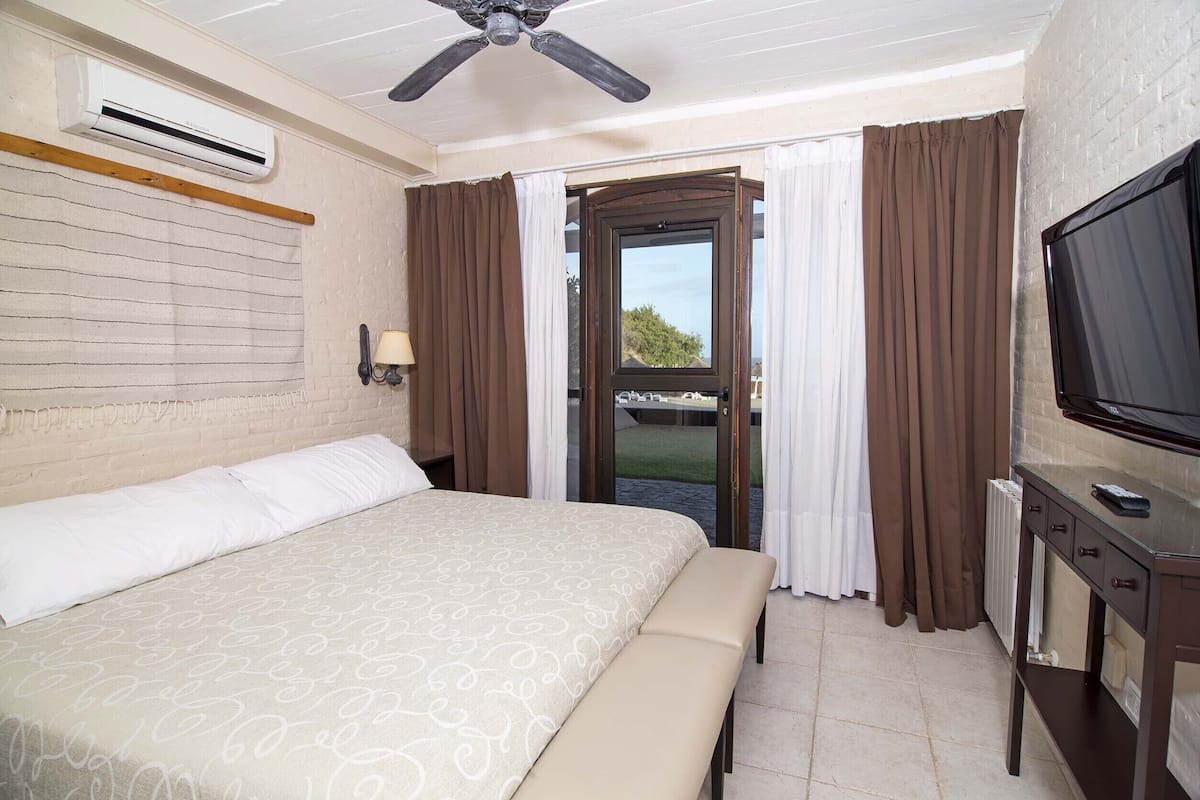 A hotel room with a bed, a bench, a TV, an air conditioner, and a view through a glass door.