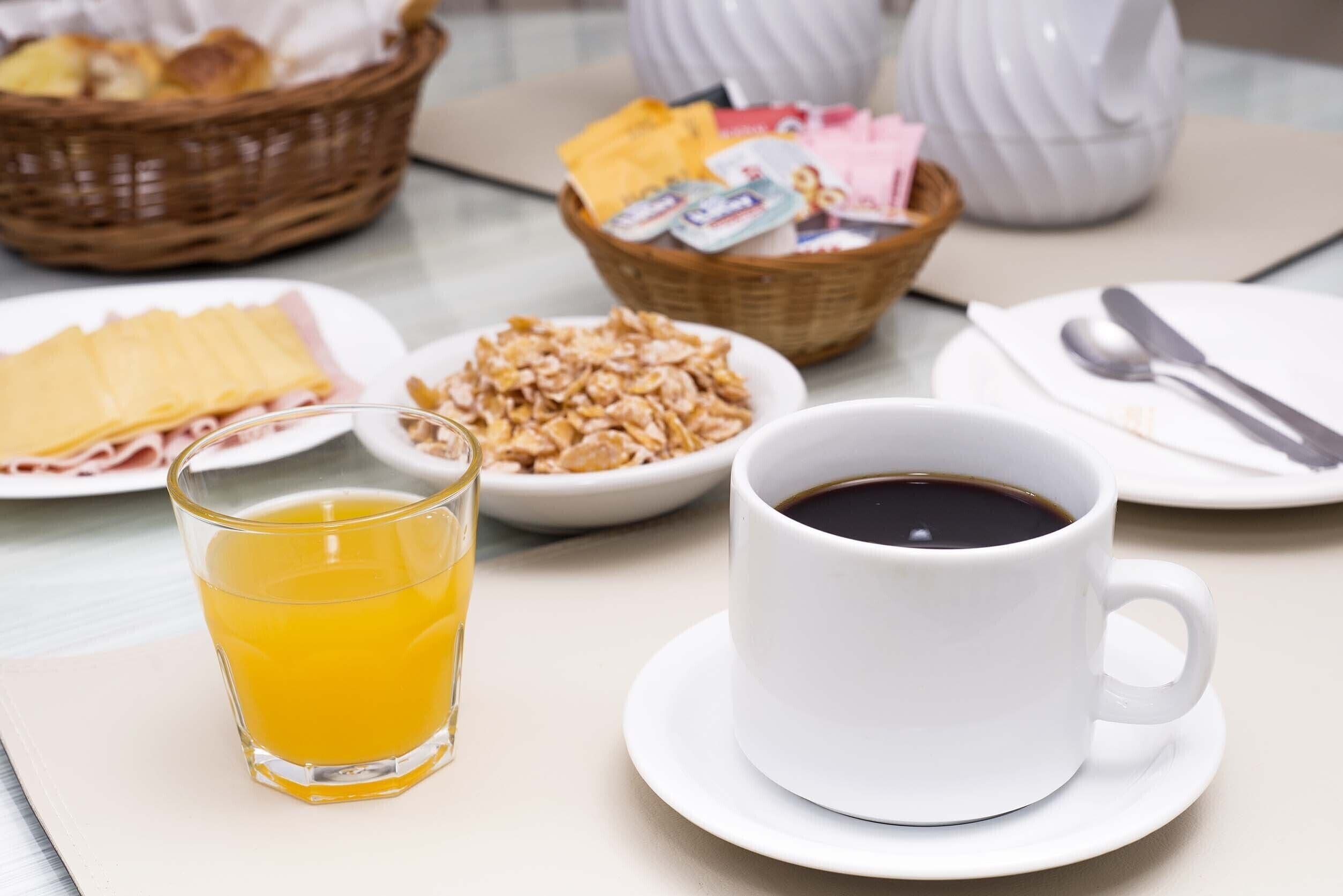 Een ontbijtsetting met een glas sinaasappelsap, een kop koffie, een bord met ham, een kom cornflakes en een mand met brood.