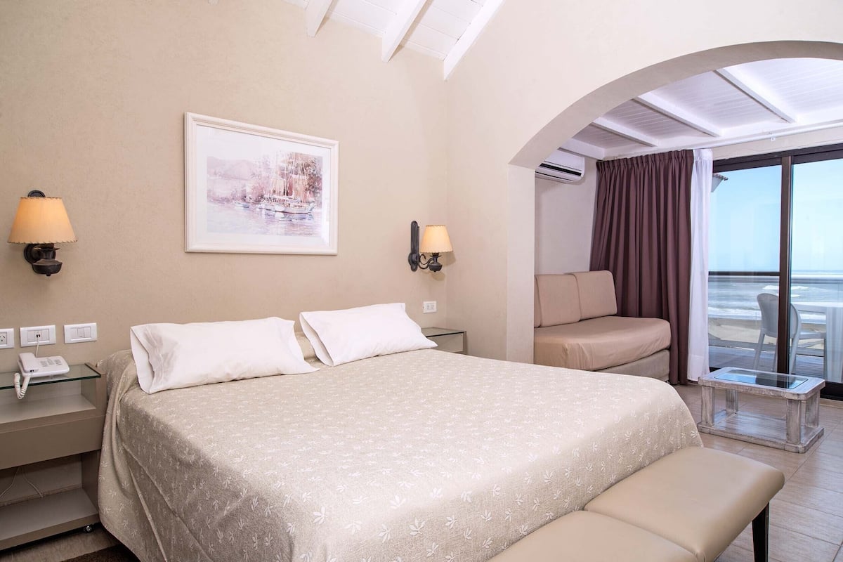 A hotel room with a bed, bedside lamps, a small table, a bench, a sofa, and a large window with curtains.