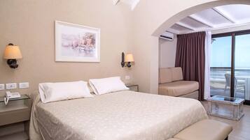 A hotel room with a bed, bedside lamps, a small table, a bench, a sofa, and a large window with curtains.