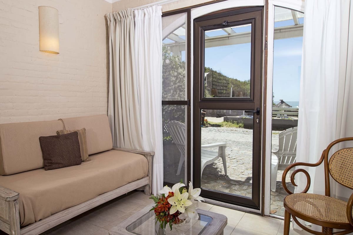 A room with a sofa, a glass coffee table with flowers, a wicker chair, and a sliding glass door leading to an outdoor area with chairs and a view of the sea.
