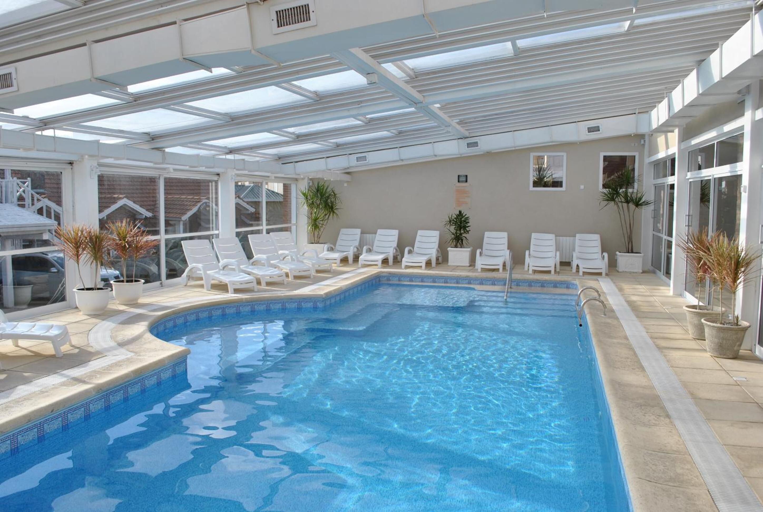 An indoor swimming pool with clear blue water, surrounded by white lounge chairs and potted plants.