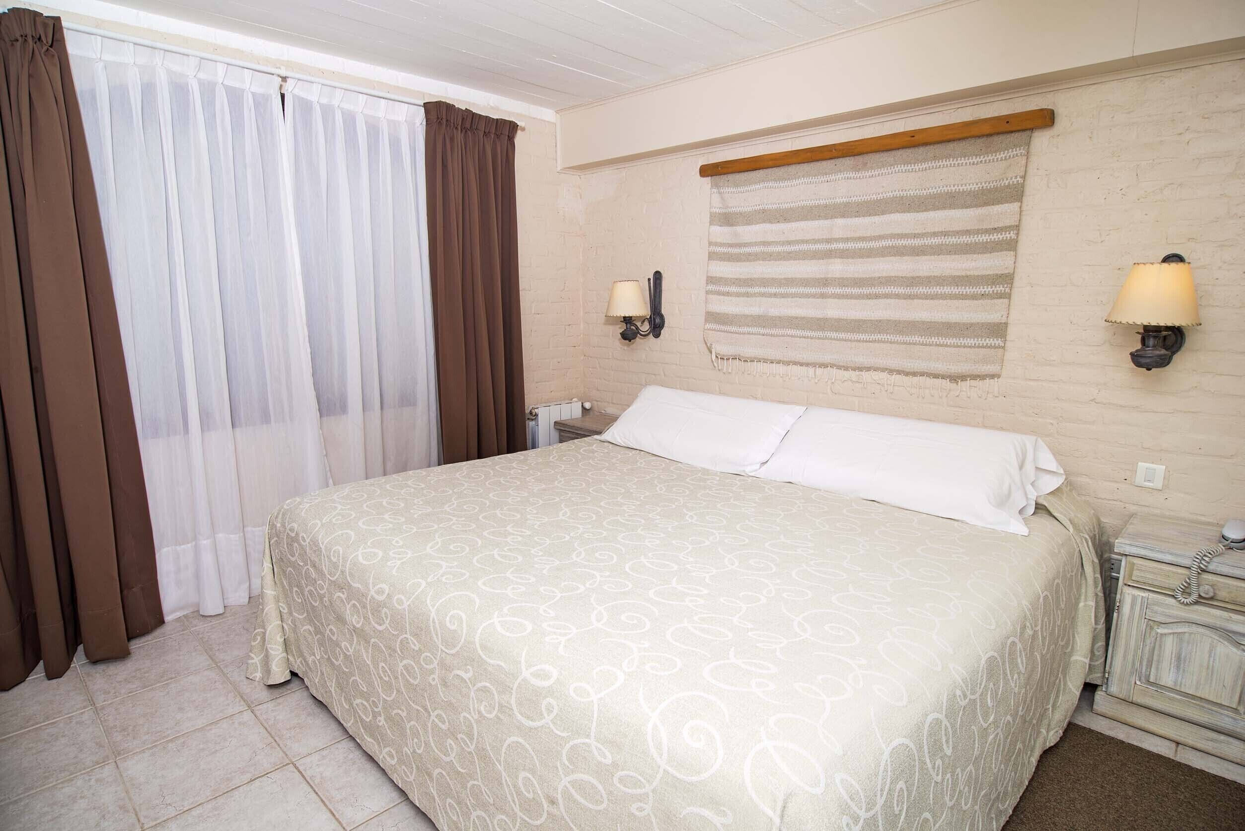 Habitación de hotel con una cama grande, cortinas, una mesita de noche, una lámpara y un teléfono instalado en la pared.