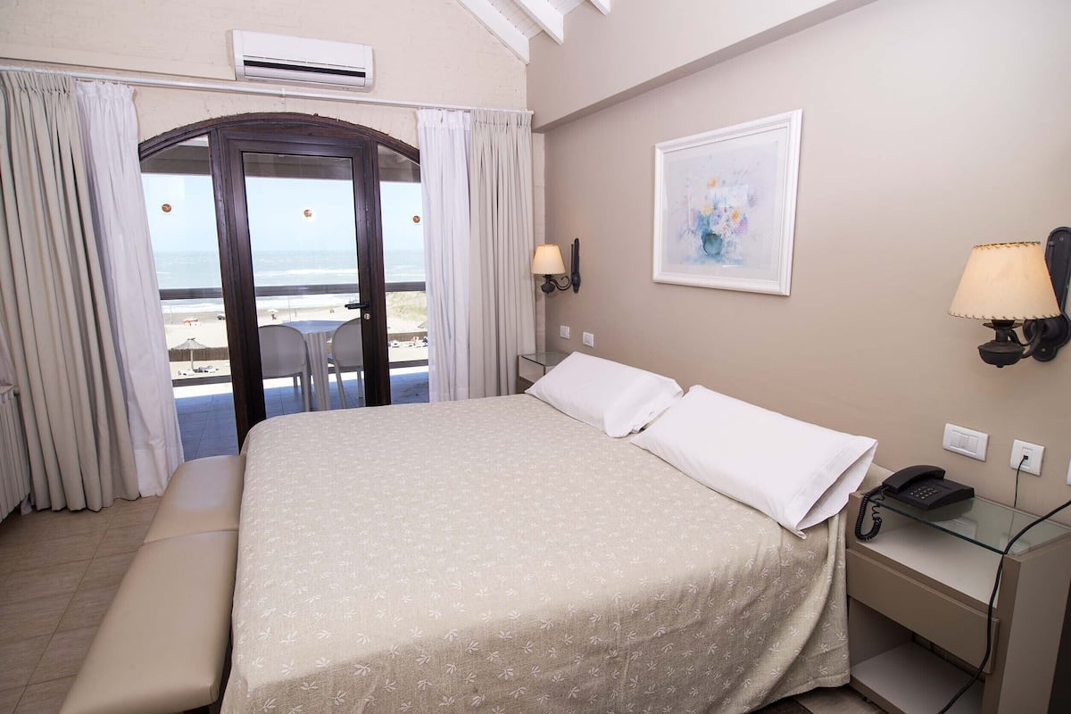 A hotel room with a bed, a desk with a phone, a lamp, a painting, and a view of the beach.