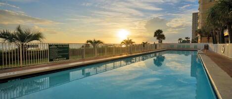 A swimming pool with a sunset view, surrounded by a fence and palm trees.