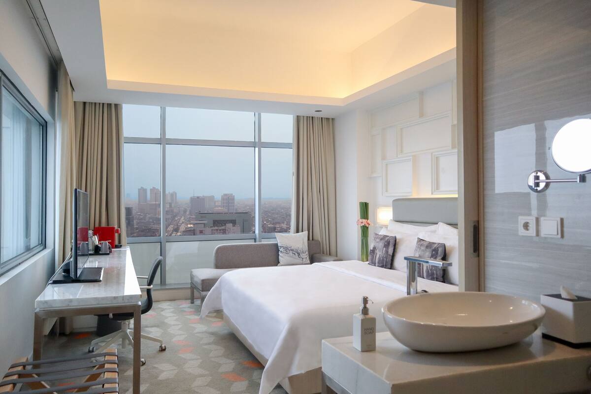 Une chambre d’hôtel avec un grand lit, un bureau avec un ordinateur, une chaise, un grand miroir rond et une vue sur la ville grâce à de larges fenêtres.