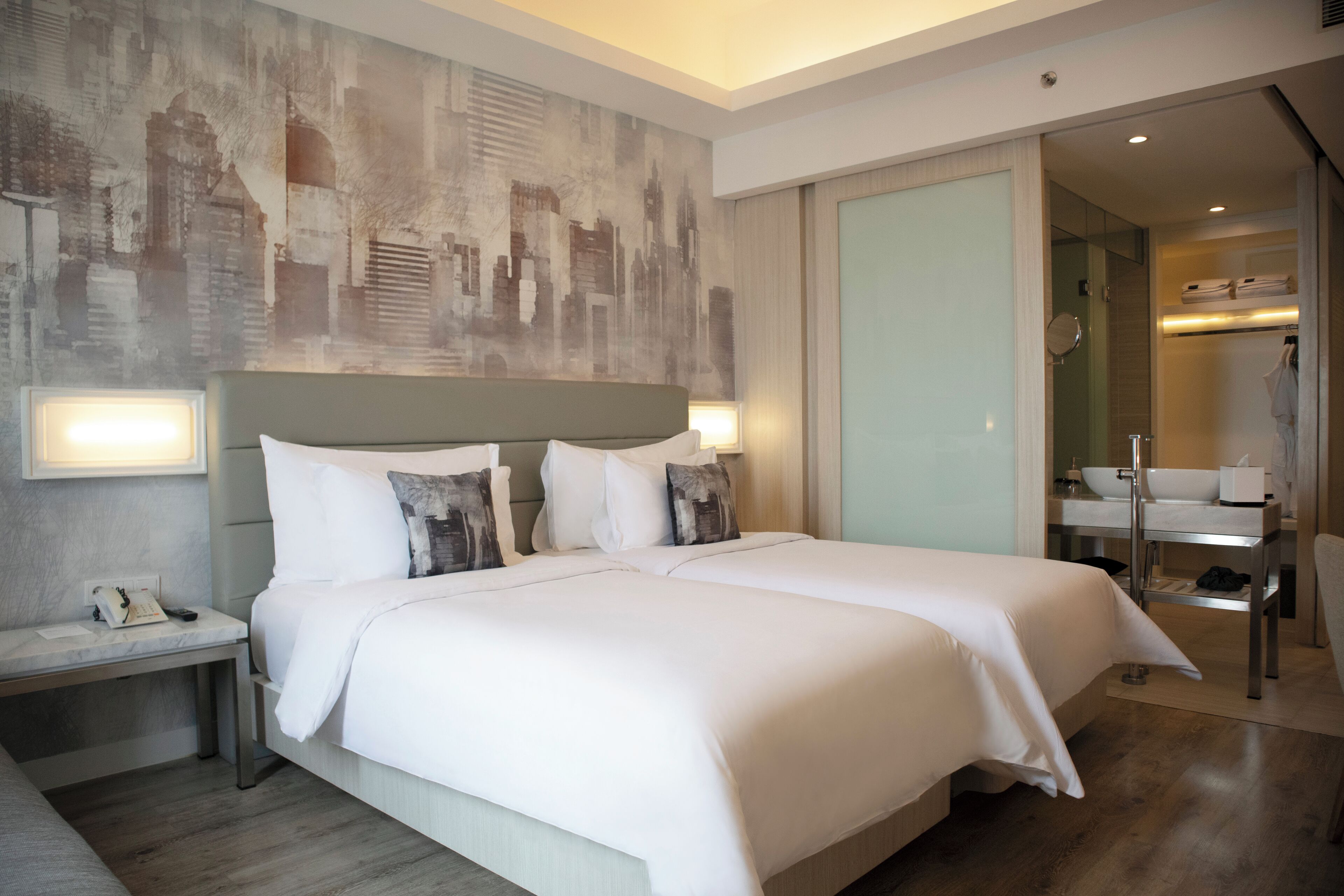 Habitación de hotel con dos camas, una mesita de noche, baño con lavamanos y espejo, y un mural con vista a la ciudad.