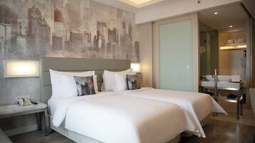 Habitación de hotel con dos camas, una mesita de noche, baño con lavamanos y espejo, y un mural con vista a la ciudad.