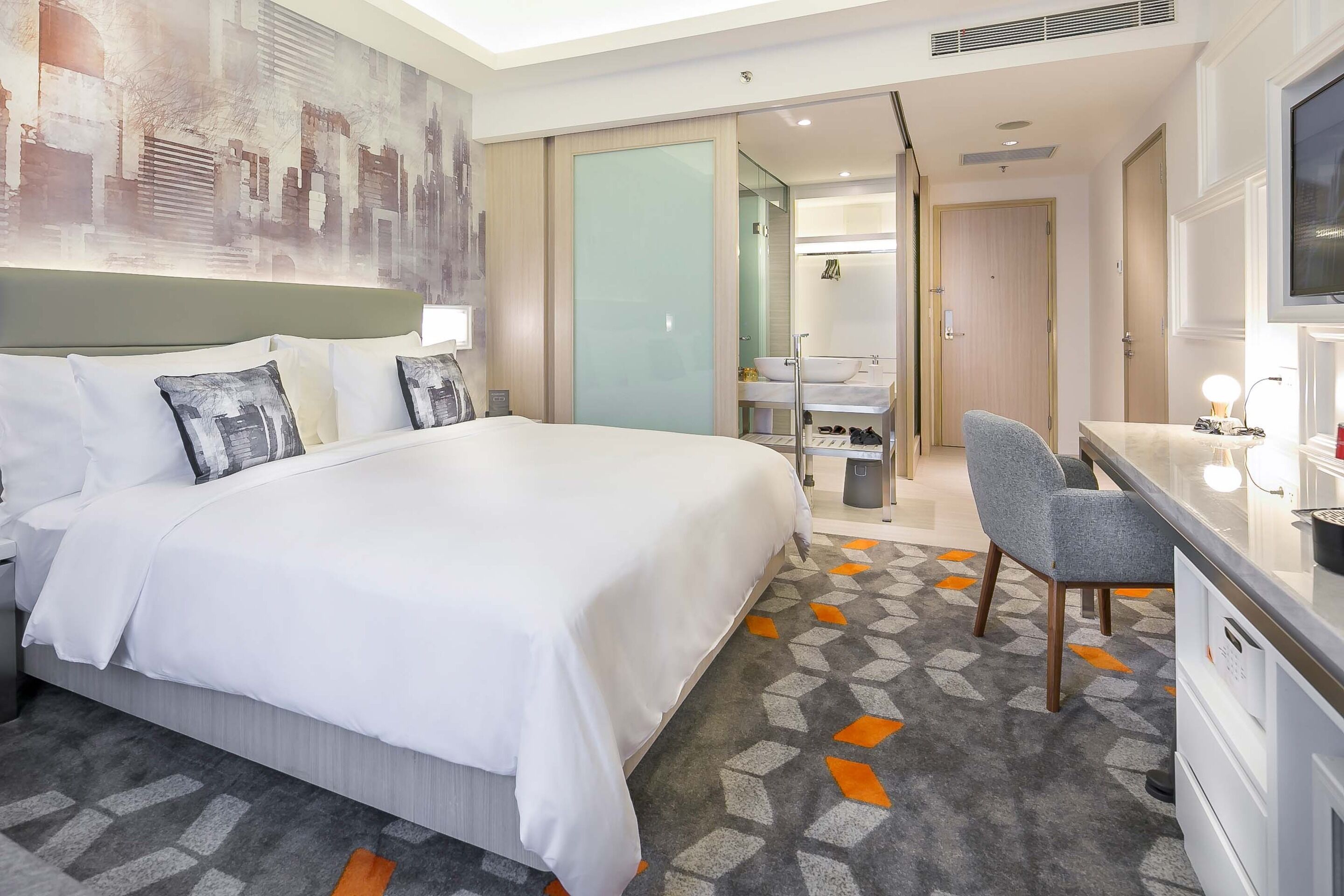 A hotel room with a large bed, a desk with a chair, a bathroom, and a cityscape artwork on the wall.
