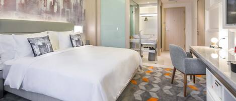 A hotel room with a large bed, a desk with a chair, a bathroom, and a cityscape artwork on the wall.
