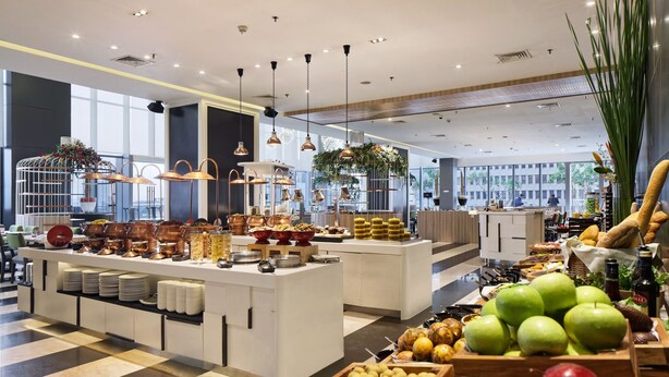 Un moderno vestíbulo de hotel con una zona de bufé que ofrece diversas frutas y pasteles.