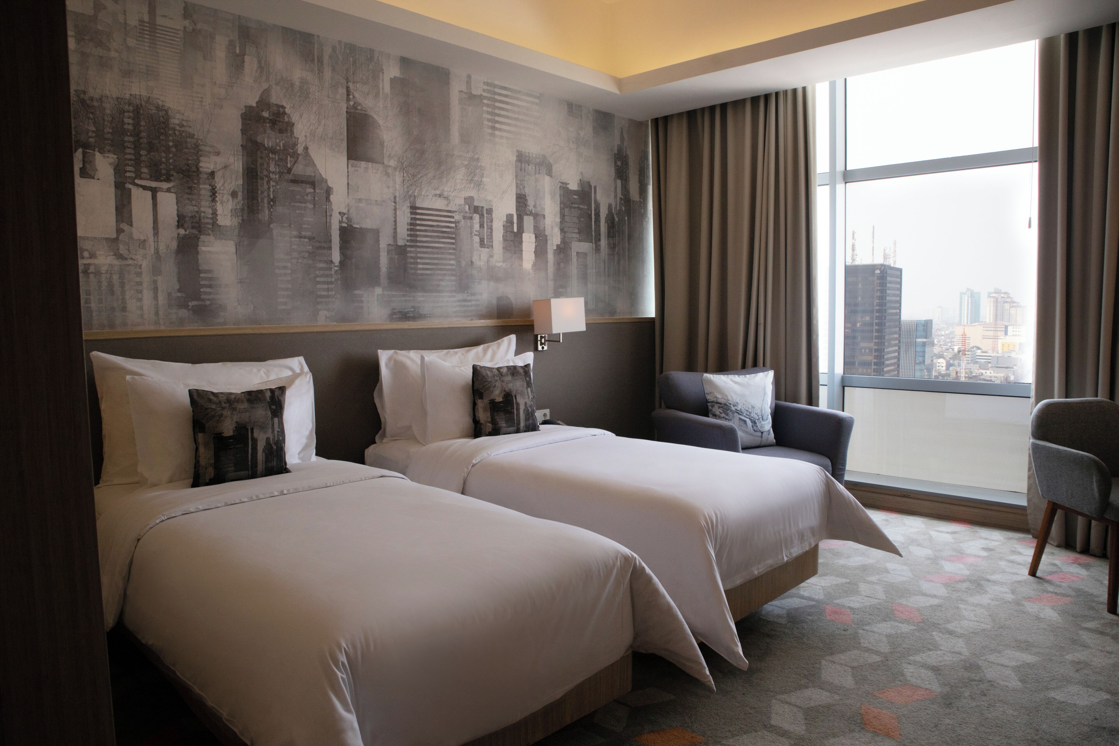 Habitación de hotel con dos camas, un mural de paisaje urbano en la pared, un ventanal con cortinas y vistas al horizonte de la ciudad.