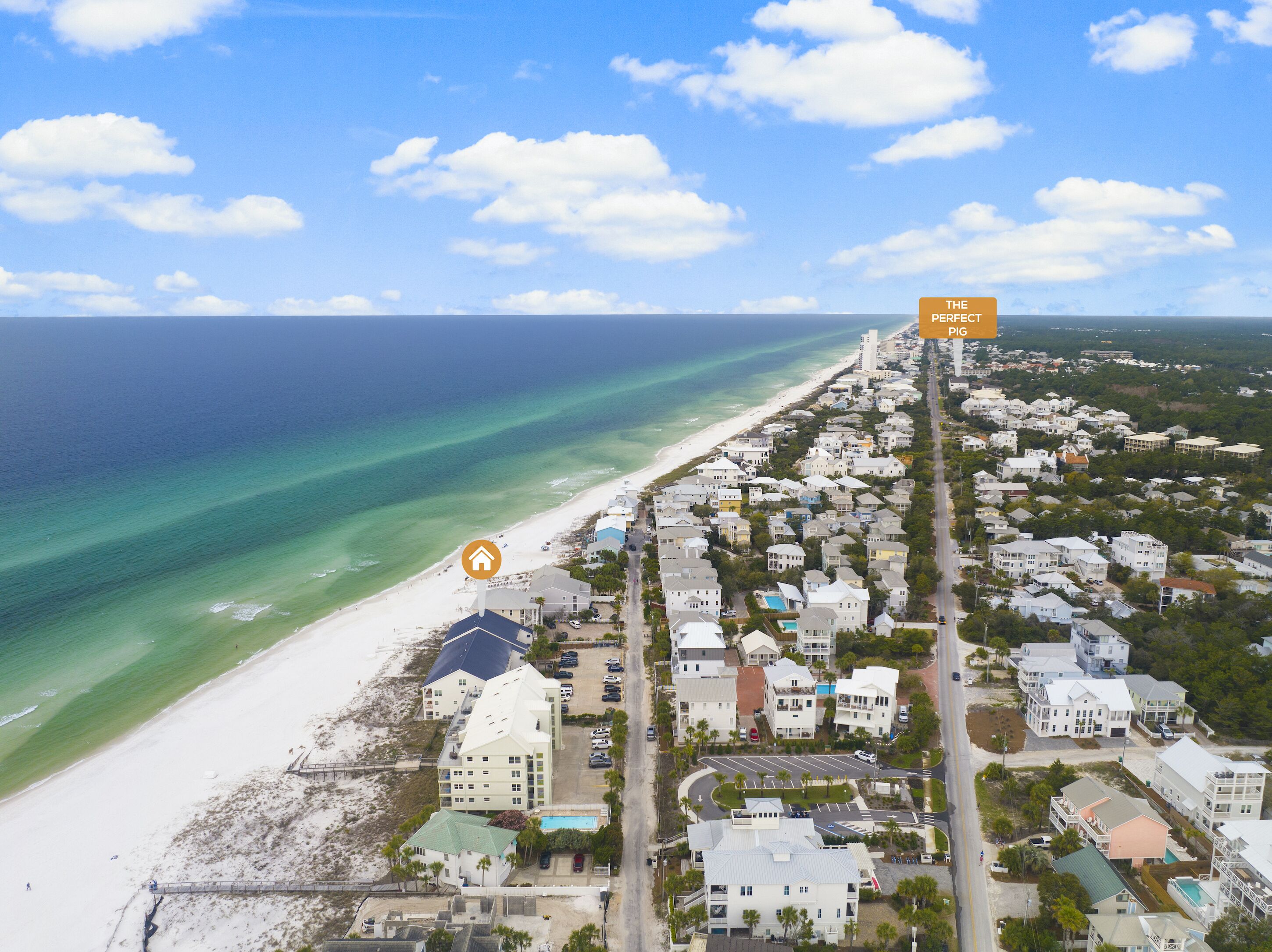 An aerial view of a coastal town with a beach, houses, and a prominent hotel with a large pool.